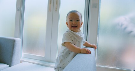 Curious Asian Baby Exploring Home Environment on Sofa in Bright Living Room with Natural Light with a curious expression.