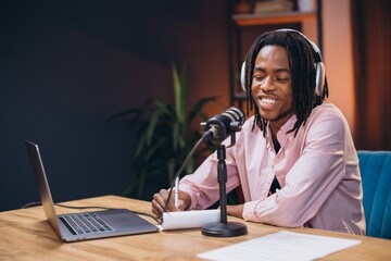 Young African American man wearing headphones and using a microphone, laptop and notepad, recording audio content for a podcast