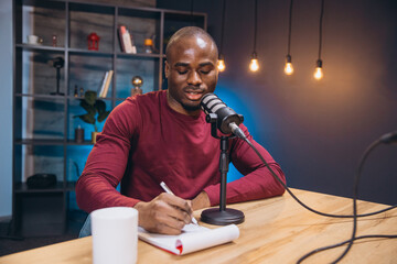 Young African American podcaster speaking into microphone and taking notes while recording podcast...