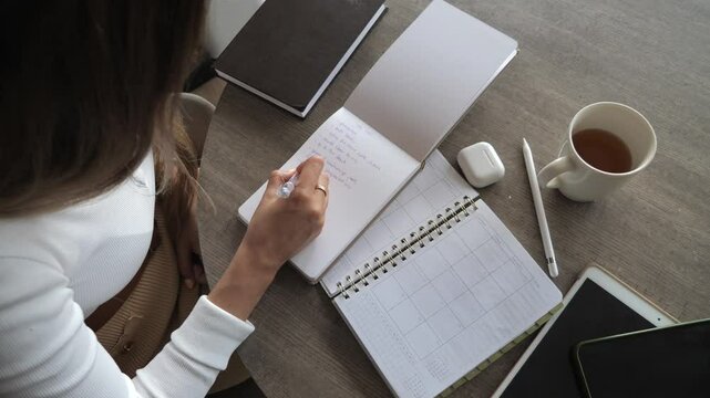 Woman writing to-do list in notebook with coffee, planner, and gadgets. Productivity, time management, and organization concept.