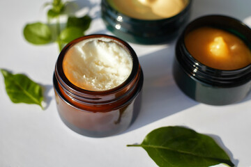 Close-up of natural body butters and balms in glass jars with green leaves around. Organic skincare concept in sunlight.