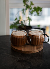 two glass mugs filled with frothy coffee sit on a wooden tray atop a dark marble surface, with a blurred plant in the background adding a cozy, relaxed atmosphere. rounded, light, soft, setting