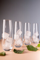 four tall transparent glasses arranged in a line, each containing pristine ice cubes, placed on a smooth beige surface adorned with scattered fresh green leaves. balanced, tranquil, calm, organized
