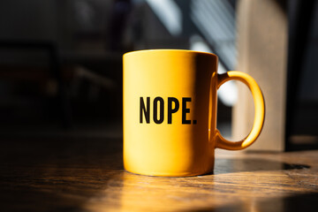 Yellow coffee mug with bold black text reading “NOPE.” sits on a wooden surface, partially lit by natural sunlight.
