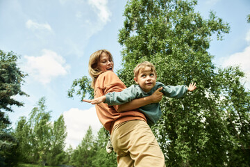 Fototapeta premium Low angle view at young mother spinning son flying like airplane in green park, enjoying time outdoors