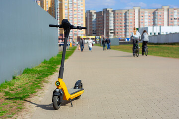A bright yellow electric scooter stands on a paved pathway beside a metal fence.