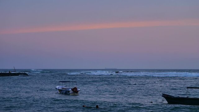 Peaceful scene of a calm ocean at sunset, with pastel colors in the sky, small boats floating on the water, and swimmers enjoying the tranquil sea. Gentle waves roll in the background, creating a rela