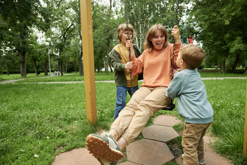 Portrait of happy family with two children at playground in park, with two boys pushing mom on swing and having fun together