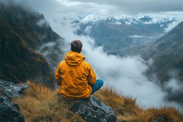 A man sits meditating on a mountain peak, overlooking a valley shrouded in clouds and snow-capped peaks
