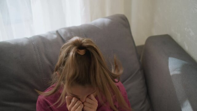 Close-up of little child with red bangle and tied hair sneezing into tissue while resting on couch due to flu, child looks unwell and exhausted with natural light filtering through the window