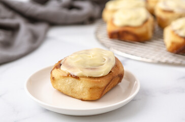 Homemade GLazed Cinnamon Roll on a White Plate and White Marble Kitchen Counter; Additional Rolls on Cooling Rack in Background