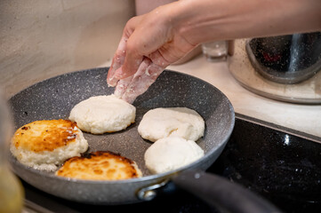 Frying Syrniki – Female Hands Covered in Dough and Flour. Close-up of female hands frying traditional syrniki (cottage cheese pancakes) in a frying pan.