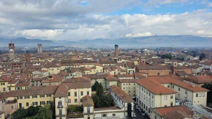 Obraz premium Panoramic view of a historic Italian town of Lucca with red-tiled roofs, towers, and mountains in the distance under a cloudy sky.