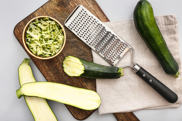 Wooden board with fresh green zucchini and bowl of slices on grey background