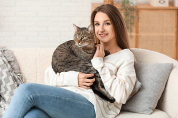 Pretty young woman holding cute tabby cat sitting on sofa at home
