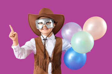 Funny little boy in cowboy costume and carnival mask pointing at something on purple background
