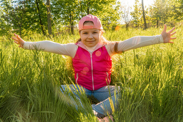 A cheerful girl sits in the thick green grass and spreads her arms to the sides. Walk outdoors in the city park