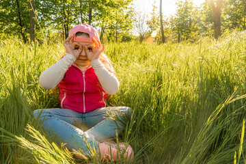 A cheerful girl sits in the thick green grass and puts her palms to her face in the form of glasses. Walk outdoors in the city park