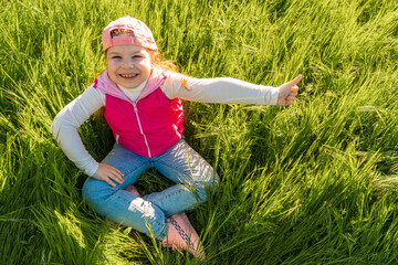 A cheerful girl sits in the thick green grass and makes a hand gesture with a finger raised up. Walk outdoors in the city park