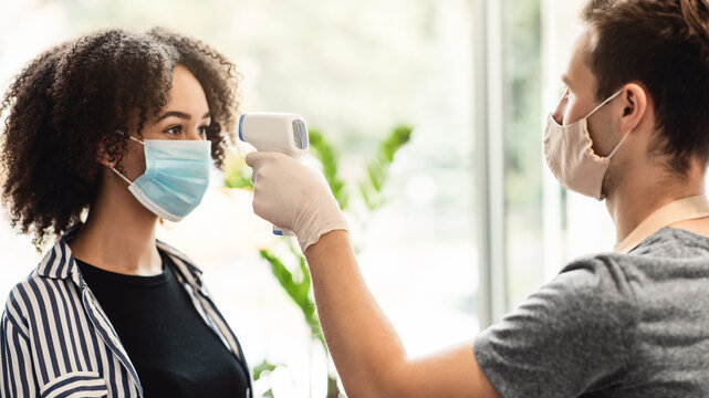 Young manager in protective mask and gloves checking temperature on client forehead at beauty salon, side view - Powered by Adobe
