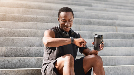 Smiling African Athete Guy Checking Fitness Tracker After Training Outdoors, Sitting On Urban Steps, Copy Space