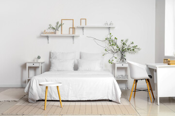 Vase with blooming branches on bedside table near cozy bed in white bedroom