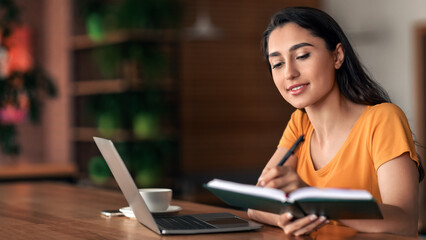Beautiful young woman planning her day, writing in notepad, sitting in front of laptop, drinking coffee or tea, cafe interior, side view, empty space. Pretty arab lady taking notes while websurfing