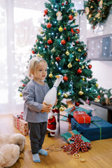 Little smiling girl stands with a toy duck in her hands near a decorated Christmas tree with gifts