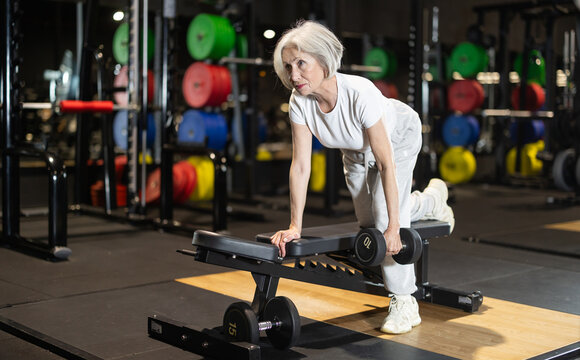 Senior woman performing one-arm dumbbell row on flat bench, using strength training to support posture, back muscles, and overall mobility. Concept of healthy lifestyle of older generation.. - Powered by Adobe