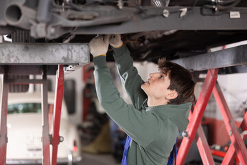 Young guy mechanic in uniform repairs underbody of car in car service station