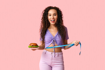 Happy young African-American woman with measuring tape and burger on pink background. Diet concept