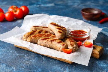 Baking paper with tasty grilled sausages, tomatoes and bowl of sauce on blue background