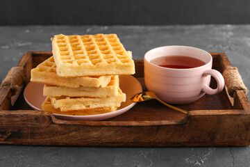 Wooden tray with tasty Belgian waffles and cup of tea on black background