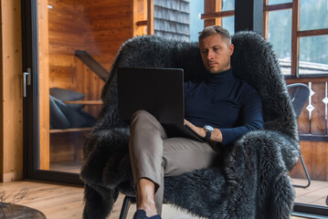 A man in a dark turtleneck and beige pants sits in a fur covered chair, using a laptop. Large...
