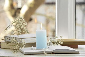 Burning candle with books and flowers near window, closeup