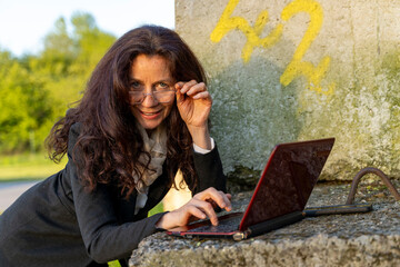 beautiful middle-aged businesswoman typing on laptop leaning on concrete bollard as outdoor desk