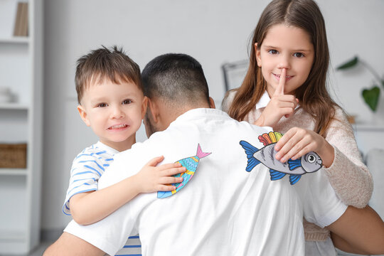 Little children attaching paper fishes to their father's back and showing silent gesture at home. April fool's day prank