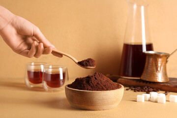 Female hand with wooden bowl of coffee powder, spoon, sugar cubes, glasses and cezve on beige background