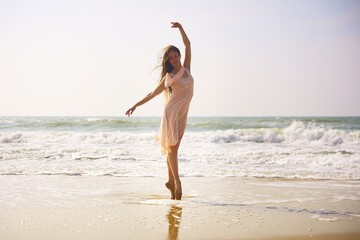 A ballet dancer stands en pointe on wet sand as waves roll behind her. Dressed in a light flowing dress, she strikes a graceful pose beneath the gentle light of a quiet seaside morning. 