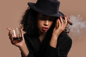 Sexy African-American woman with whiskey smoking cigar on brown background