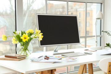 Vase with flowers and blank computer monitor on desk near window in office