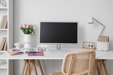 Vase with tulips, magazines and blank computer monitor on desk in light office