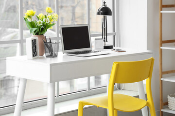 Blank laptop, clock and vase with spring flowers on desk near window in office