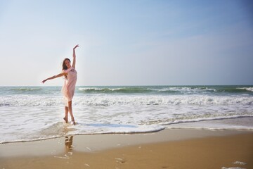 A graceful ballet dancer performs en pointe at the water edge, wearing a flowing pink dress. Her elegant pose contrasts beautifully with the foamy waves and wide open blue sky above the sea.