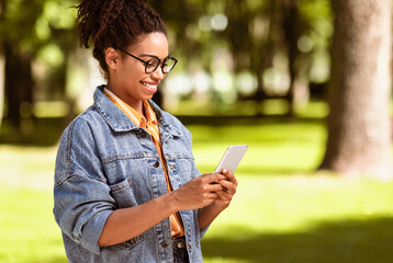 Happy Black Woman Holding Smartphone Reading Good News Message Walking Outside In Park. Mobile Application Concept. Copy Space