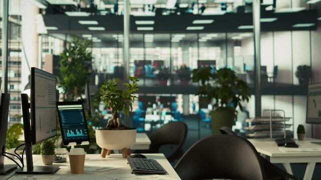 Business graphs and performance metrics on monitors in workplace, handheld camera shot. Office desk setup in bright corporate environment with monitors displaying data analytics, panning shot