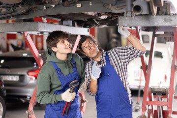 Young guy and adult male mechanic in uniform repairing underbody of car in car service station