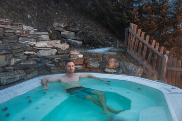 A man sits in a hot tub surrounded by a stone wall and wooden fence, with evergreen trees, a rocky hillside, and snow patches in a mountain setting.