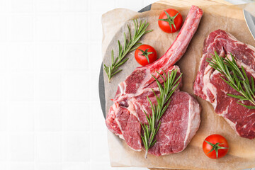 Pieces of raw beef, rosemary and tomatoes on white tiled table, top view. Space for text