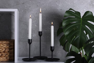 Stylish candlesticks with burning candles on shelf and houseplant near grey wall indoors